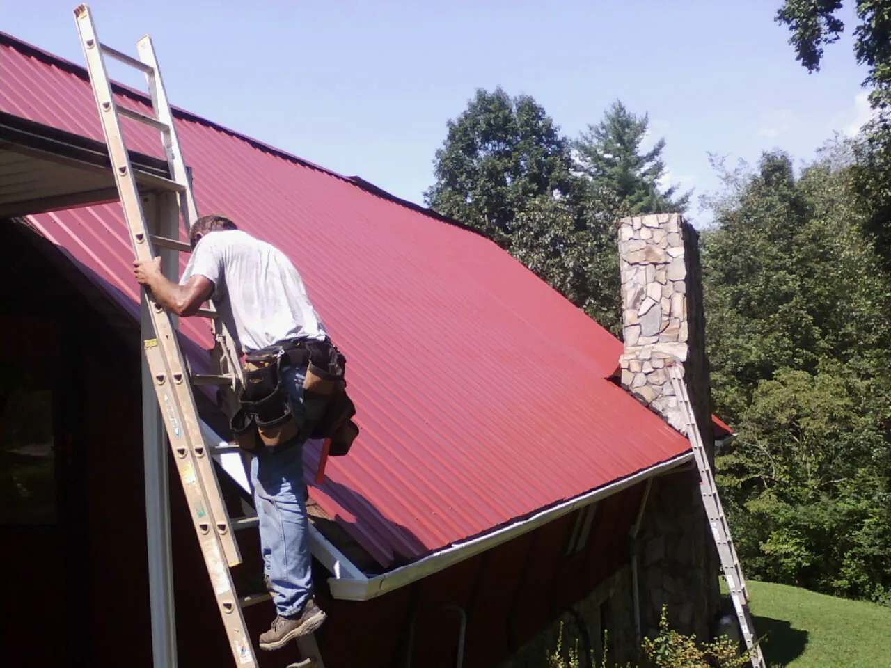 Roofer on a ladder beside a red metal roof and stone chimney — Soffit & Fascia Repair by Roofing Brothers in Wilmore